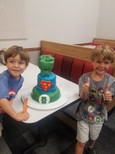 Two kids on the sides of a three tiered superhero cake, with Green lantern on the bottom, superman in the middle, and hulk's fist at the top.